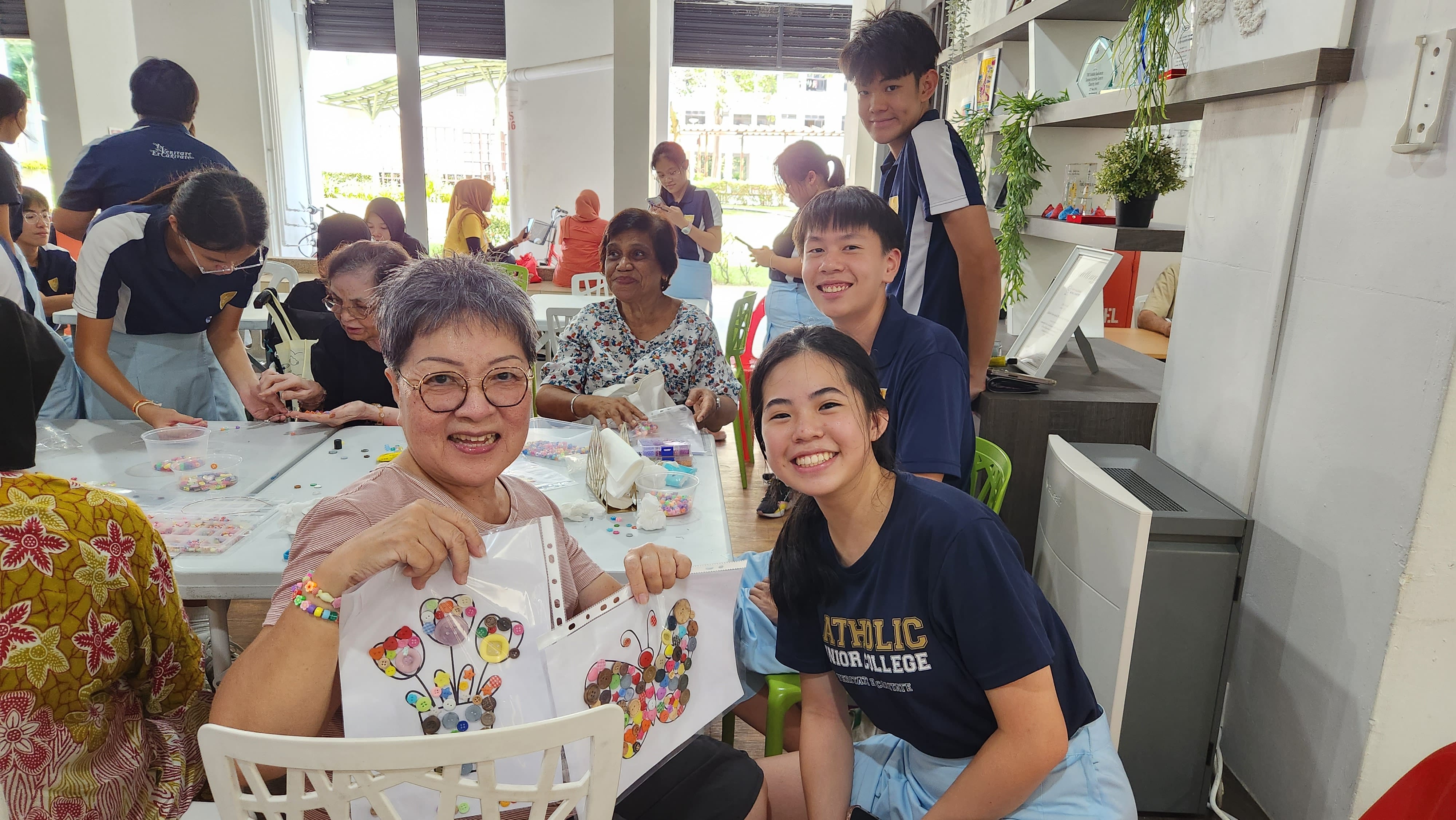 An elderly woman and students in navy uniforms smile holding butterfly button art crafts at a community activity table.